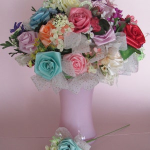 May include: A bouquet of silk flowers in a pink vase. The bouquet includes roses in shades of pink, blue, and red, as well as white and cream flowers. The bouquet is decorated with white tulle and lace.