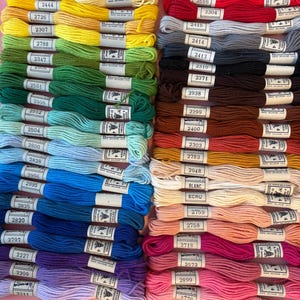 May include: A collection of colorful embroidery floss skeins, arranged in neat rows. The floss is in a variety of shades, including red, orange, yellow, green, blue, and purple. Each skein has a label with a number. The image is well-lit and shows the texture of the thread.