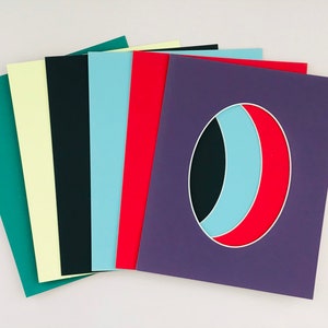 May include: A set of eight different colored cardstock sheets. The sheets are arranged in a fan shape, with the top sheet being a purple sheet with a white oval cutout. The oval cutout has a black, light blue, and red design inside.