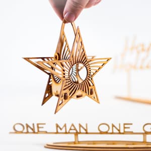 May include: A wooden star ornament with a cutout design, hanging from a string. The star is being held by a hand. The ornament is in front of a wooden sign that says "Happy Holidays". Below the star is another wooden sign that says "One Man One Garage".