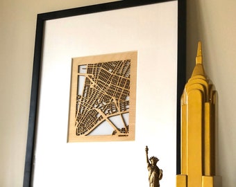 May include: A framed wooden map of a city with a black frame, a gold statue of the Statue of Liberty, and a yellow ceramic building on a white mantel.