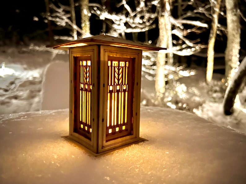 May include: A wooden lantern with a geometric design and a warm glow, sitting on a snowy surface.