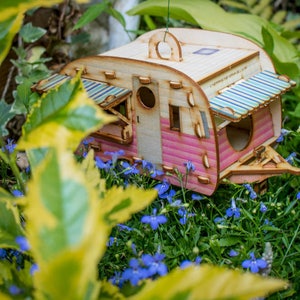 May include: A pink and white wooden birdhouse shaped like a vintage camper with a striped awning. The birdhouse is surrounded by blue flowers and green foliage.