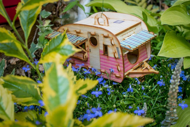 May include: A pink and white wooden birdhouse shaped like a vintage camper with a striped awning. The birdhouse is surrounded by blue flowers and green foliage.