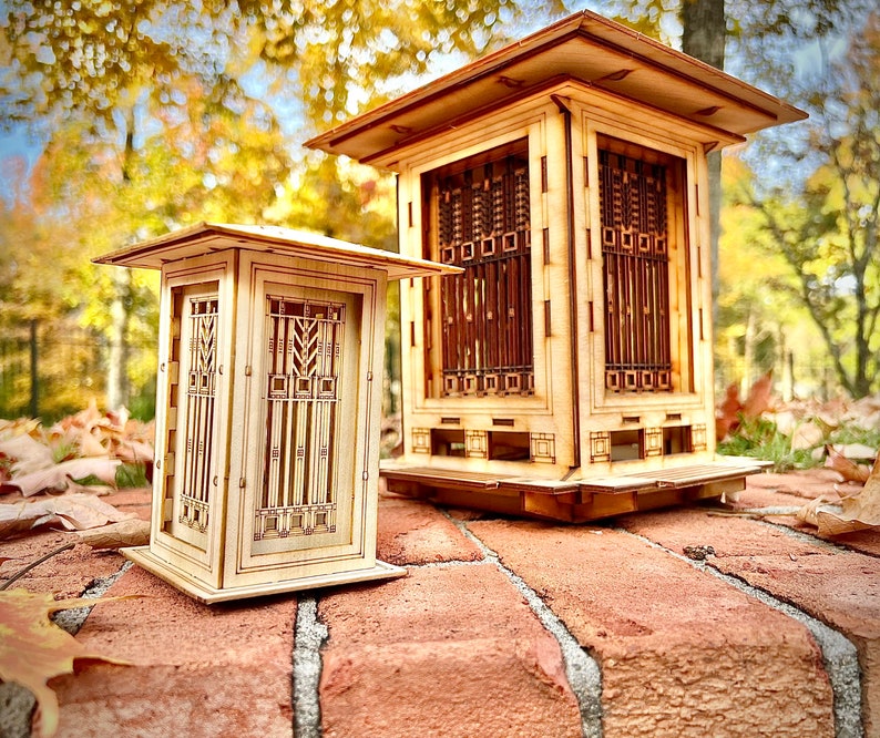 May include: Two wooden model houses with intricate window designs. The larger house has a square base and a pitched roof. The smaller house has a rectangular base and a flat roof. Both houses are made of light wood and have a natural finish.