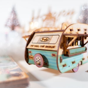 May include: A wooden model of a vintage camper van with a blue and white paint scheme. The camper has a sign that reads "One Man, One Garage".
