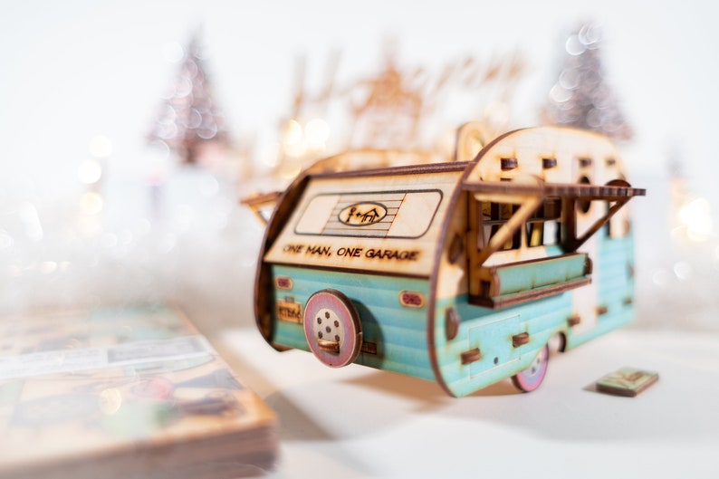 May include: A wooden model of a vintage camper van with a blue and white paint scheme. The camper has a sign that reads "One Man, One Garage".