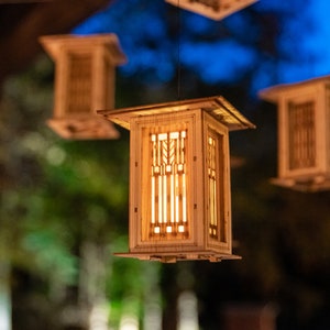 May include: Four wooden hanging lanterns with geometric designs and warm light shining through. The lanterns are suspended from the ceiling against a blurred background of trees and a blue sky.