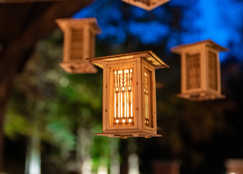 May include: Four wooden hanging lanterns with geometric designs and warm light shining through. The lanterns are suspended from the ceiling against a blurred background of trees and a blue sky.