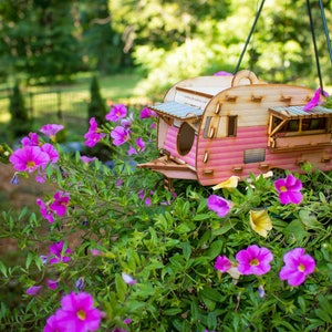 May include: A pink and white wooden birdhouse shaped like a vintage camper trailer hanging from a branch in a garden with pink flowers.