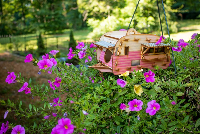 May include: A pink and white wooden birdhouse shaped like a vintage camper trailer hanging from a branch in a garden with pink flowers.