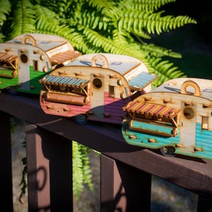 May include: Three miniature wooden camper vans in different colors, green, pink, and blue, with striped awnings and windows. The campers are lined up on a wooden railing.
