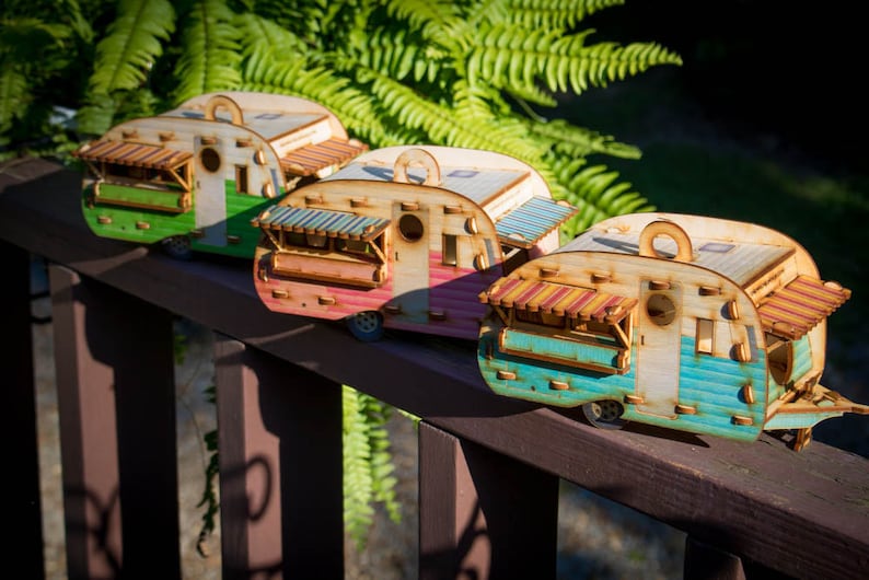 May include: Three miniature wooden camper vans in different colors, green, pink, and blue, with striped awnings and windows. The campers are lined up on a wooden railing.