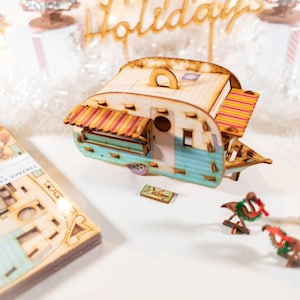 May include: A wooden model of a vintage camper with a blue and white exterior, a red and yellow awning, and a wooden sign that says "Welcome". The camper is surrounded by wooden decorations, including a flamingo with a Christmas wreath, a grill, and a sign that says "Holidays".