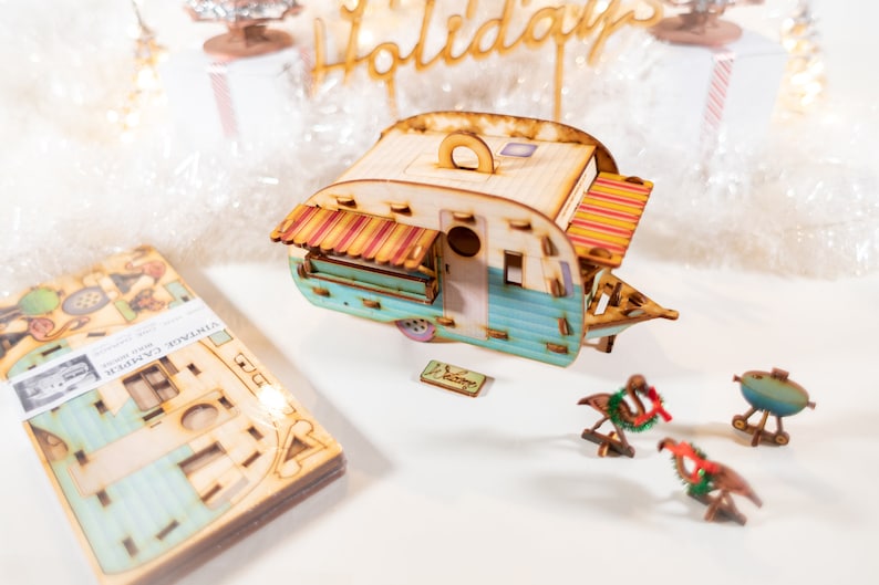 May include: A wooden model of a vintage camper with a blue and white exterior, a red and yellow awning, and a wooden sign that says "Welcome". The camper is surrounded by wooden decorations, including a flamingo with a Christmas wreath, a grill, and a sign that says "Holidays".