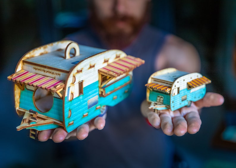 May include: Two wooden model camper vans with a teal blue body, white roof, and red and white striped awnings. The larger camper van has the text "CREATIVEMONEYTRADE.COM" on the side.