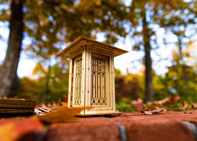 May include: Detailed close-up of a miniature wooden model of a Prairie School-style structure. The model is light brown, exhibiting intricate geometric patterns and a gabled roof.  The design features vertical lines and geometric window details. The model is situated outdoors on a brick surface, with blurred autumn foliage in the background, suggesting a fall setting. The craftsmanship is evident in the fine details of the miniature building.