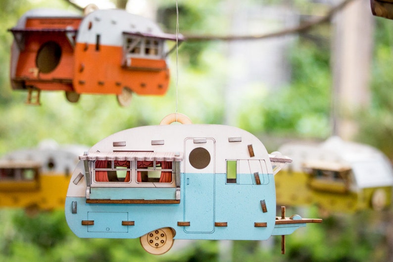 May include: Three wooden birdhouses shaped like vintage campers. The birdhouses are painted in various colors, including blue, white, orange, and yellow. The birdhouses have small windows and doors, and they are hanging from a tree branch.