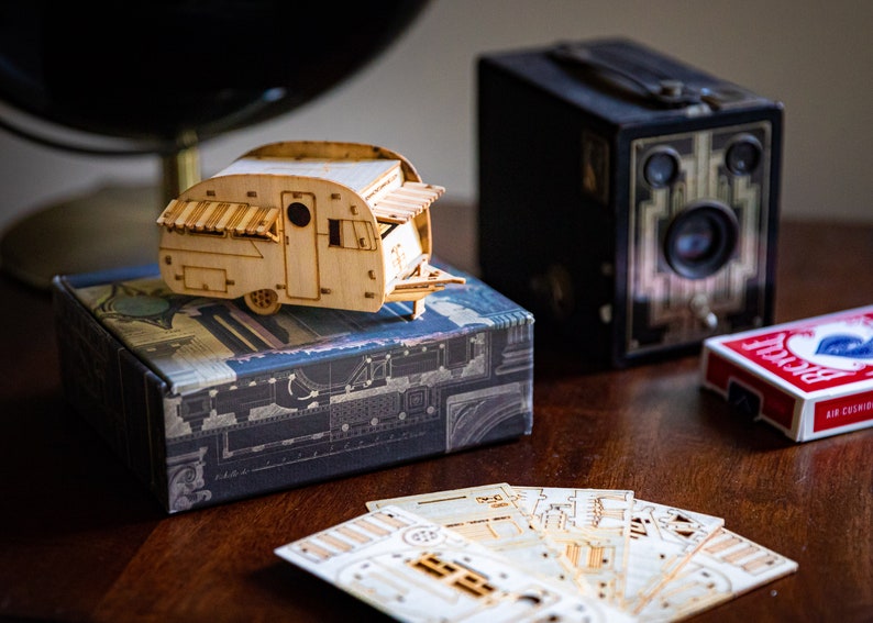 May include: A wooden model of a vintage camper trailer with a striped awning, sitting on a box with a patterned design. There are also a deck of cards and an old camera in the image.