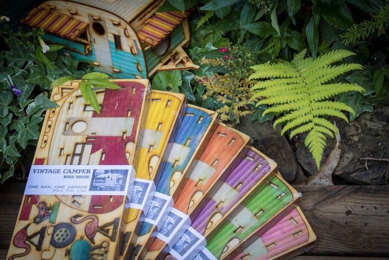 May include: A set of eight colorful wooden birdhouses shaped like vintage campers. Each birdhouse has a different color scheme and features a cutout design. The birdhouses are labeled "Vintage Camper Bird House" and "One Man, One Garage, One Play, One Love".