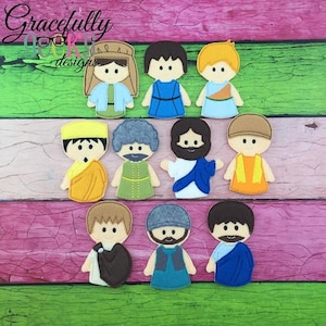 May include: A collection of felt figures depicting biblical characters. Each figure has a unique outfit and hairstyle, with colors including blue, yellow, brown, and green. The figures are arranged on a pink and green wooden surface.