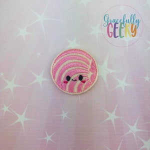 May include: A circular embroidered patch featuring a smiling, cartoon-style pastry with pink and white stripes. The patch has a cream-colored border and black eyes. The background is a light pink with white star shapes, and the text "Gracefully GEEKY" is in a circle.