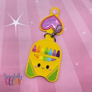 May include: A yellow crayon box keyring with a smiling face and a pink heart charm. The keyring features a rainbow of crayons inside the box.
