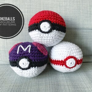 May include: Three crocheted Pokeball amigurumi toys. The Pokeballs are red, white, and purple with black accents. The purple Pokeball has a white "M" on it. The text "POKEBALLS AMIGURUMI PDF PATTERNS" is in a black circle.