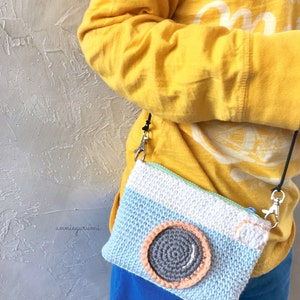 May include: A light blue crocheted camera bag with a gray and orange lens. The bag has a zipper closure and a black strap. The bag is being worn by a person wearing a yellow shirt.