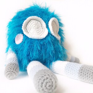 May include: A blue and gray crocheted plush toy with white eyes and pink cheeks. The toy is sitting on a white surface.