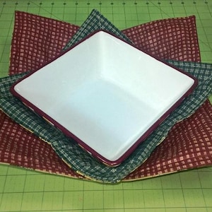 May include: A white ceramic bowl with a red rim sits inside two fabric squares. The squares are green and white plaid and red and white plaid.