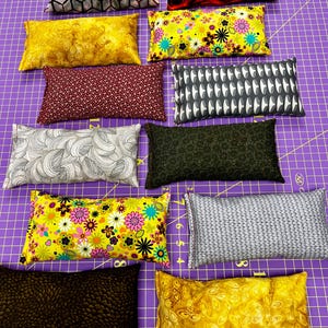 以下が含まれることがあります： さまざまな模様と色を持つ長方形の布製枕のコレクション。デザインには、黄色、赤、黒、グレー、緑の色合いのフローラル、幾何学模様、抽象模様が含まれます。枕は紫色のカッティングマットに配置されています。