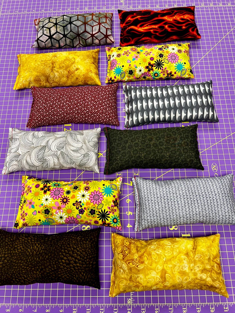 May include: A collection of rectangular fabric pillows with various patterns and colors. Designs include floral, geometric, and abstract patterns in shades of yellow, red, black, gray, and green. The pillows are arranged on a purple cutting mat.
