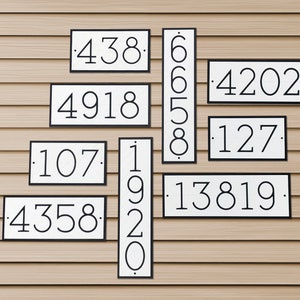 Multiple house number signs of the same style are mounted to a large wall of vinyl siding. The sizes of the plaques are different, as are the quantity of numbers on each plaque. A variety of signs with different digits and orientations for reference.