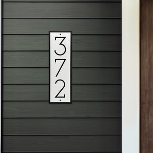 The house number sign is shown from a distance on a home with siding. The numbers on the plaque are clearly visible and are a modern farmhouse style. The proportions of the address sign look appropriate for the home.