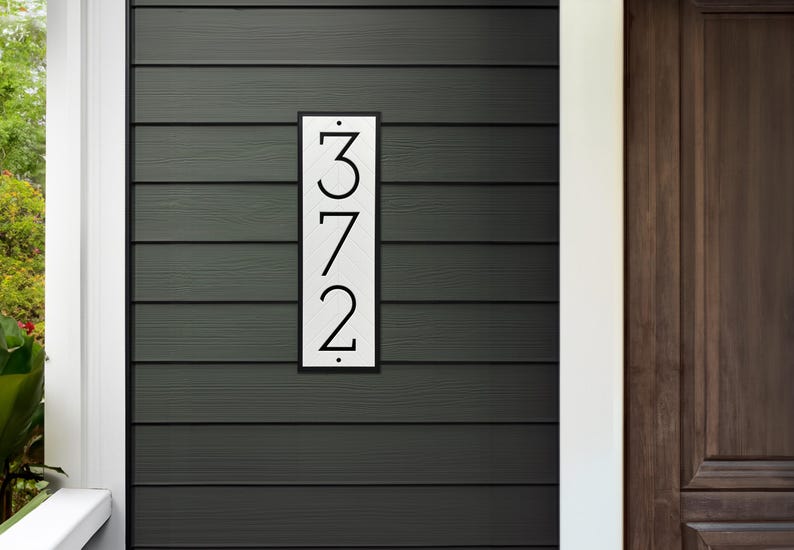 The house number sign is shown from a distance on a home with siding. The numbers on the plaque are clearly visible and are a modern farmhouse style. The proportions of the address sign look appropriate for the home.