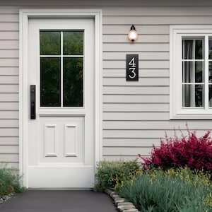 The house number sign is shown from a distance on a house with vinyl siding. The numbers on the plaque are clearly visible and the proportions look appropriate for the home. Shrubs and a stone pathway make this a beautiful scene around the sign.