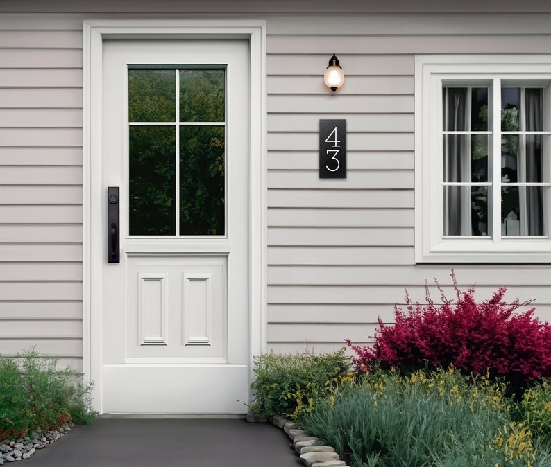 The house number sign is shown from a distance on a house with vinyl siding. The numbers on the plaque are clearly visible and the proportions look appropriate for the home. Shrubs and a stone pathway make this a beautiful scene around the sign.