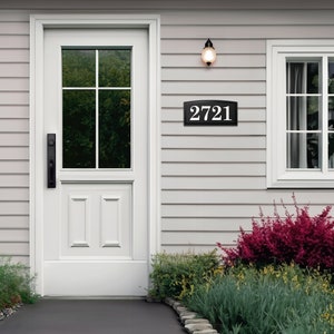 The house number sign is shown from a distance on a house with vinyl siding. The numbers on the plaque are clearly visible and the proportions look appropriate for the home. Shrubs and a stone pathway make this a beautiful scene around the sign.