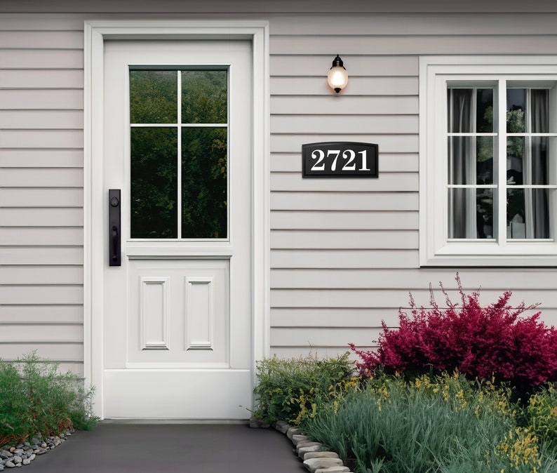 The house number sign is shown from a distance on a house with vinyl siding. The numbers on the plaque are clearly visible and the proportions look appropriate for the home. Shrubs and a stone pathway make this a beautiful scene around the sign.