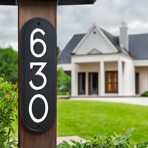 May include: Black and white house number plaque with the numbers 630 on a wooden post. The plaque is oval shaped and has a modern design.
