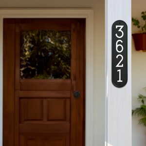 May include: Black vertical house number plaque with white numbers 3621. The plaque is mounted on a white wooden post.