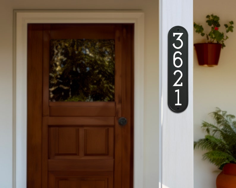May include: Black vertical house number plaque with white numbers 3621. The plaque is mounted on a white wooden post.
