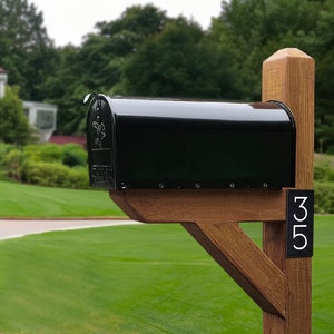 May include: A black mailbox with a wooden post and a house number plaque that reads "353". The mailbox is mounted on a wooden bracket attached to the post. The mailbox is in front of a green lawn and a house in the background.
