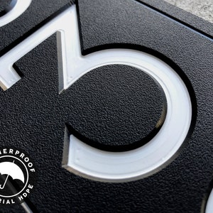 May include: Close-up of a black house number sign with white raised numbers "3" and "0". The sign is made of weatherproof high-density polyethylene (HDPE) material.