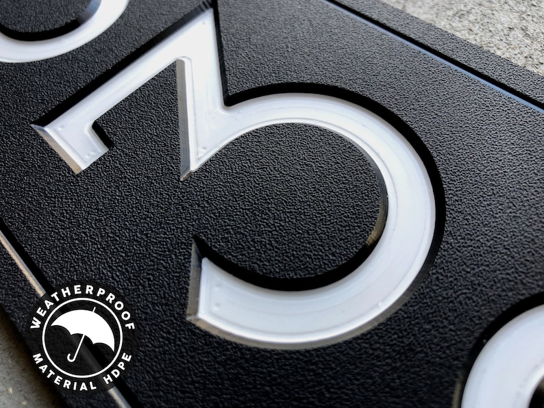 May include: Close-up of a black house number sign with white raised numbers "3" and "0". The sign is made of weatherproof high-density polyethylene (HDPE) material.