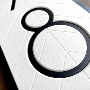 close up view that shows the details of the sign. The black, modern numbers are recessed into the white geometric background. There is a clean chamfered edge around the profile of the numbers. The quality looks professional and precise.