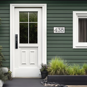 The house number sign is shown from a distance on a house with vinyl siding. The numbers on the plaque are clearly visible and the proportions look appropriate for the home. Shrubs and a stone pathway make this a beautiful scene around the sign.