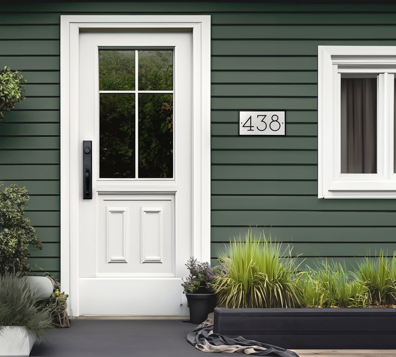 The house number sign is shown from a distance on a house with vinyl siding. The numbers on the plaque are clearly visible and the proportions look appropriate for the home. Shrubs and a stone pathway make this a beautiful scene around the sign.