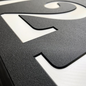 Close up view of the house number plaque. The surface of the sign is textured. The white numbers are recessed and are cleanly cut into the sign. Crisp lines show the quality of the work on the final product.